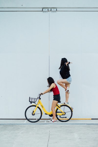 一名女子骑自行车，另一名女子站在靠墙的后挡泥板上